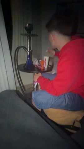 A young man in a red hoodie and denim shorts sits cross-legged, using a cell phone next to a hookah on a small, black table. A Burger King cup and various small objects sit on the table. He exhales a large cloud of smoke. The scene is dimly lit, with light from a window covered by sheer curtains. A blurred painting hangs on the wall behind him. The bottom half of another person is visible in the foreground, implying they are seated.