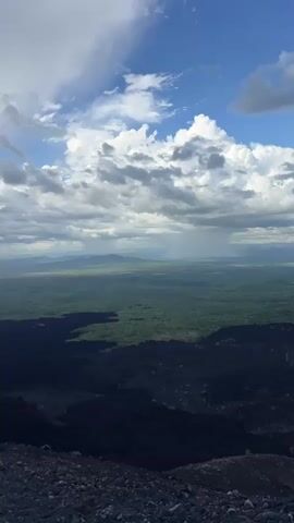 This media file shows a panoramic view of a volcanic landscape. The foreground is dominated by dark, rocky terrain, characteristic of volcanic ash and lava fields. In the middle ground, the landscape transitions to lush green hills and a vast expanse of vegetation, suggesting a fertile area adjacent to the volcanic zone. In the background, distant mountains are visible under a dramatic sky.

The sky is filled with a mixture of light and dark clouds, with some areas showing sunlight breaking through, creating rays of light. There are indications of rain in the distance, with visible streaks of precipitation falling over the green landscape and mountains.

The time of day appears to be daytime, likely late afternoon or early evening, given the quality of the light and the presence of dramatic cloud formations. The weather seems to be dynamic, with sunshine and rain occurring simultaneously in different parts of the scene.

The overall scene evokes a sense of raw, natural power and the contrast between barren volcanic land and life-sustaining greenery. The elevated perspective suggests the footage is filmed from a high vantage point, possibly the rim of a volcano or a nearby peak. There is no visible text in the media file.
