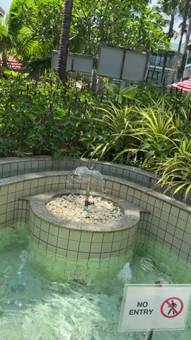 The video shows a small ornamental fountain with a gentle stream of water flowing through it. The fountain is located in an outdoor area with lush green foliage and palm trees. In the background, there is a multi-story building that appears to be a hotel or resort. The water in the fountain is clear and shallow, with small white pebbles at the bottom. There is a "NO ENTRY" sign with a symbol of a person being crossed out, indicating that no one is allowed to enter the fountain. A person wearing a light-colored t-shirt and shorts is walking away from the fountain on a wooden walkway. The sky is blue with a few white clouds.