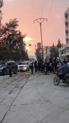 The video depicts a street scene at sunset. Several pedestrians and motorcyclists are visible on a cracked road. Cars are parked and driving slowly. In the distance, a building with a "ROSE" sign is visible. Police lights are flashing on top of a vehicle in the middle of the group.  A power pole stands tall, with wires stretching across the sky. Trees line the street, decorated with small flags. The sky is a mix of orange and pink hues.