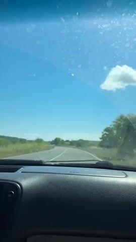 This is a first-person perspective video taken from inside a car driving on a paved road. The dashboard of the car is visible in the foreground, with various buttons and a car stereo. The windshield shows a bright blue sky with a few fluffy white clouds. The road ahead is curving to the left, with grassy fields and trees on either side. The sun appears to be shining, casting a warm light on the landscape. There are no other vehicles or people visible in the immediate vicinity. The video appears to be shot during the daytime.