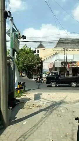 A black pickup truck drives through an intersection. A person on a motorbike with a large white box on the back drives through the intersection. A person on a bicycle rides through the intersection. The sky is blue with white clouds. There are buildings and trees in the background. Power lines are visible. The ground is paved.