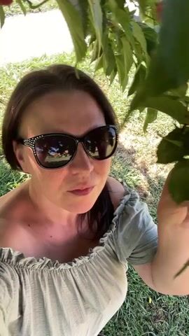 A woman wearing sunglasses and an off-the-shoulder top is sitting outdoors under a tree. She is holding something small in her fingers and bringing it to her mouth. The background consists of green grass and leaves.