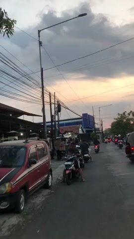 The video depicts a street scene with several people and vehicles, likely in a busy urban or semi-urban area. The time of day appears to be late afternoon or early evening, as indicated by the lighting and the sky, which shows hues of orange and yellow mixed with clouds.

In the foreground, a red car is parked on the left side of the frame. Further down the street, several motorbikes are visible. One motorbike in the center of the frame has a person sitting on it, facing towards the camera. Another motorbike is positioned just behind it.

On the right side of the street, there are people gathered around a food cart or stall. This cart appears to be mobile, attached to a motorbike or a similar vehicle. A person wearing a helmet and a dark t-shirt is standing near the cart. To their left, another person in a light-colored shirt and grey pants is also present. Further to the right, a person in a pink headscarf and blue jeans is visible.

In the background, there are buildings, including what appears to be a mosque with a distinctive roof and a blue building with signage. Electrical wires are strung across poles overhead, which is a common sight in many urban environments. The overall impression is of a bustling street with everyday activity, possibly a market area or a place where people gather to eat.