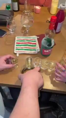 Here is a detailed description of the video:

A tabletop is laden with bottles of various alcoholic beverages, including Get 27 liqueur and a bottle of gin, alongside shot glasses containing clear liquid. Two individuals are visible, each arranging the shot glasses. One person, wearing a blue and green striped shirt, carefully places a shot glass on the table. The setting appears to be indoors, with a domestic environment indicated by the kitchen counter in the background. A striped potholder, a box of projector lights, and other miscellaneous objects are scattered around the tabletop, suggesting a casual, social gathering.