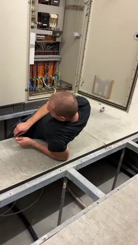 In a dimly lit, indoor industrial setting, likely a factory or control room in Mannheim, Germany, two individuals are engaged in electrical work. The primary focus is on a large, open electrical cabinet filled with various wires and components. The cabinet doors are a neutral beige color, and the interior is a chaotic yet organized arrangement of orange, blue, and other colored wires, along with circuit breakers and terminal blocks.

The first individual, seen in the foreground from a slightly elevated perspective, is a bald man wearing a black t-shirt and dark pants. He is positioned on a raised platform, leaning into the open floor space beneath the electrical cabinet, working with his hands. His posture suggests he is focused on a task within the wiring system, possibly making adjustments or connections.

A second individual, a man wearing a navy blue t-shirt and dark blue work pants, is more prominently featured in the latter half of the video. He is kneeling in front of the electrical cabinet, his back to the camera. He is actively interacting with the cabinet, his hands reaching inside, presumably performing electrical maintenance or installation. His t-shirt appears damp, indicating physical exertion.

Surrounding the individuals are various tools and equipment. In the background, a metallic toolbox with an assortment of screwdrivers and other tools is visible. A red and white striped traffic cone stands nearby, and several spools of white electrical wire are scattered on the floor. A few electrical plugs and connectors are also present. The overall scene suggests a professional environment where electrical systems are being maintained or commissioned. The lighting is utilitarian, typical of an industrial space, and there are no indications of weather or time of day beyond the artificial illumination. The emotions conveyed are focused and professional, with no overt displays of stress or urgency. No visible text is present in the media.