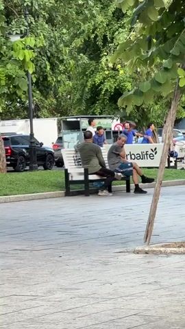 The video shows an outdoor scene with several people and objects. In the foreground, a paved area is visible, with trees and foliage in the background. On a bench, two men are seated, facing away from the camera. One man wears a green shirt and dark pants, while the other wears a grey shirt and patterned shorts. Behind them, a white truck with a green and white logo, possibly a food truck, is parked. Several other individuals are standing near the truck and around the bench. A black SUV is parked on the left side of the frame. Towards the right, a tree trunk is prominent, and a motorcycle with a rider wearing a helmet is moving towards the camera, slightly blurring the background. The overall environment appears to be a public outdoor space, possibly a park or street with a seating area.