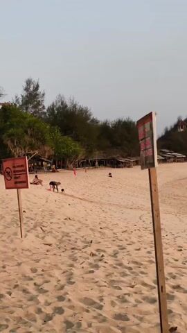A beach scene with a sandy foreground and trees in the background. There are several people visible on the beach. In the foreground, a person is kneeling and interacting with something on the sand. To the left, a sign with red text and a symbol is visible. To the right, a wooden post with a sign is also present. A few beach umbrellas or huts are lined up in the middle ground. The sky is clear and overcast.
