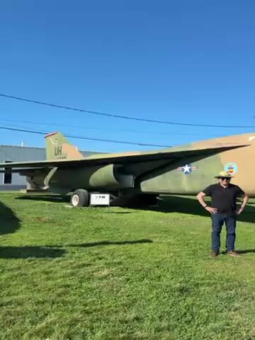 The video shows an olive drab and brown camouflage painted military jet on display on a grassy lawn under a clear blue sky. A man wearing a black shirt, jeans, and a tan hat stands beside the jet, gesturing towards it. The jet has a United States Air Force star on its side and the letters "UH" on its tail. A sign with the letters "FW" is visible near the jet's nose. The video zooms in on the front of the jet, showing a pitot tube with a yellow tip. Power lines are visible in the background.