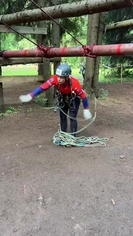 A young person wearing a helmet, a red and blue jacket, and black leggings is preparing to climb a rope obstacle course. They are wearing a safety harness with ropes attached. They are standing on a dirt ground in a wooded area. In front of them is a pile of coiled ropes. There is a wooden structure above them with ropes and poles. The person is holding a rope and appears to be adjusting it.
