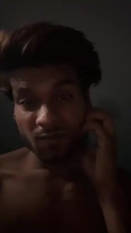 A person with dark hair and a beard, wearing no shirt, is visible from the chest up. The background is dark, with a dark gray wall and a blurred black object that may be a fan. The person is looking at the camera and touching their neck and chest. The lighting is dim.