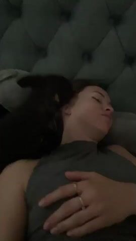 A close-up video shows a woman lying on her back in bed, propped up against a tufted headboard. A small black dog with tan markings is positioned on top of her, nuzzling her face with its snout. The woman is wearing a gray tank top and has gold rings on two fingers of her left hand, which rests on her chest. A light-colored stuffed animal is visible behind her head, and a watch is on her left wrist. The lighting is dim, suggesting it is nighttime or a darkened room.