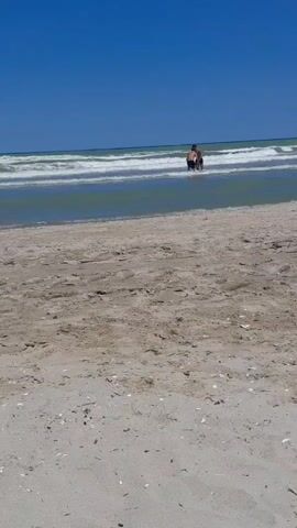 Two people are wading in the shallow ocean water near a sandy beach. The sky is clear and blue. Waves are breaking on the shore.