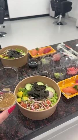 A close-up shot shows a person's hands preparing poke bowls on a countertop. In the foreground, a hand is pouring a sesame-seed dressing from a small plastic container into a poke bowl filled with rice, salmon, tuna, avocado, edamame, seaweed, and sprouts. Another poke bowl, partially visible to the left, contains similar ingredients. In the background, several small plastic containers holding ginger and wasabi, along with trays of sashimi (salmon and tuna), are arranged on the countertop. The countertop appears to be made of dark granite with red speckles. The environment seems to be indoors, possibly a kitchen or a dining area, with blurred furniture and bright lighting.