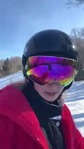 A person, possibly a young woman, is skiing down a snowy slope on a sunny day. The individual wears a red fleece jacket, a black helmet with a “uvex” logo and an American flag design, and rainbow-tinted goggles. The camera is positioned from a first-person perspective, showing the skier’s point of view. The background consists of snow-covered ground and trees, some bare and some evergreen, suggesting a forested or mountainous area. The shadows cast by the trees fall across the slope. The skier moves down the hill at a moderate pace.