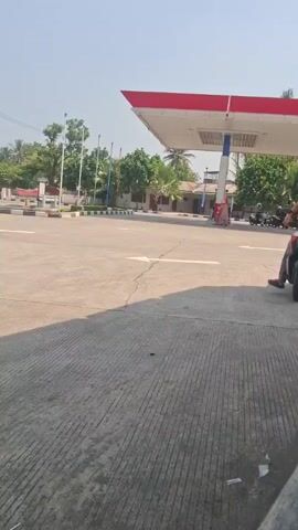 A young man wearing a brown long-sleeved shirt, gray pants with blue and white stripes, and a white helmet is sitting on a red and black scooter. He is at a gas station with a red and white canopy visible in the background. The man is looking to his right. The ground is paved concrete. There are trees and a building in the background.