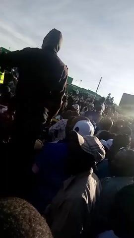 The video captures a large gathering of people outdoors under bright, sunny conditions. A prominent figure in a dark hoodie stands in the foreground, facing away from the camera, silhouetted against the bright sky. Behind them, a dense crowd of people, predominantly men, are visible. Many in the crowd wear hats or caps, and some have their hands raised in the air. A person in a red shirt is also visible within the crowd.

The atmosphere suggests a public rally or event, with collective chanting heard in the audio, including "Takbir! Allahu Akbar!". In the background, a multi-story building can be seen on the right, and a bright, clear sky dominates the upper part of the frame. The strong sunlight causes significant lens flare, particularly in the lower portion of the video. Partially visible text on a green sign in the background appears to read "Pesa", possibly "M-Pesa" or "Jum Pesa". Given the context, this event is taking place in Mwingi, Kenya, during the daytime.