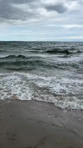 The video shows a moderately choppy ocean with waves breaking on a sandy beach. The sky is overcast with dark clouds, suggesting a stormy or impending storm. The water is dark green and appears to be turbulent. The beach is mostly empty, with some scattered debris such as seaweed and what appear to be footprints. In the distance, a few distant figures can be seen on the beach and in the water, as well as a lighthouse or tower on a hill. The camera is positioned at a low angle, looking out towards the ocean, and pans slowly to the right, revealing more of the coastline. There is no audio.