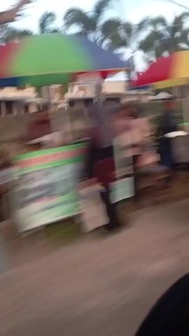 The video is a blurry, fast-moving shot of a street scene, likely taken from a moving vehicle. Several food stalls and small shops line the road. In the foreground, there's a glimpse of a motorcycle. As the view pans, a red umbrella covers a food stall with what appears to be colorful items displayed. Further down the street, other stalls with similar setups and parked motorcycles are visible. The environment suggests an outdoor market or street food area, possibly in a residential or commercial district. The lighting indicates it might be dusk or early morning. The overall impression is one of activity and commerce, though the blurriness obscures specific details of the food, people, or merchandise.