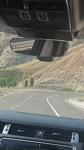 The video shows a view from inside a vehicle driving on a paved road that curves to the right. The road has white dashed lines marking the lanes and a solid white line on the right edge. On the left side of the road, there is a metal guardrail separating the road from a sloped, mountainous terrain. The terrain is covered with dry, light-brown soil, with some patches of green vegetation and trees. There are visible structures on the hills, possibly terraces or farming plots, and some utility poles. The sky is bright, suggesting daytime. The interior of the vehicle is visible in the foreground, including the dashboard, rearview mirror, and the overhead console with buttons. The rearview mirror reflects the interior of the car and a person's head.