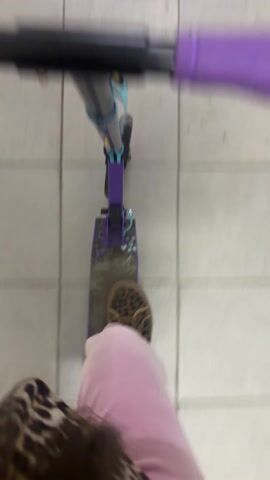 The camera, apparently attached to a person, films their point of view while riding a purple scooter. The scooter has black handlebars with purple grips, a purple frame, and what appears to be turquoise accents. The person is wearing pink sweatpants and a leopard-print headband. The environment appears to be indoors with gray and white tiled flooring, transitioning to a dark gray mat near an entrance or exit with a dark background. The camera shakes as the scooter moves.