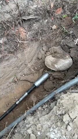 A short video clip shows a close-up view of a trench dug into the soil. Two large dark pipes are visible, one above the other, running horizontally through the trench. A metallic tool, possibly part of a drilling or digging apparatus, is inserted into the soil near the pipes. A large, light-colored rock rests on the soil in the trench, positioned above the metallic tool. The surrounding area is covered in dirt, roots, and some scattered debris, including pieces of brick. In the final moments of the clip, the view pans slightly to reveal the orange handle of a shovel partially buried in the trench. The overall environment appears to be an outdoor, possibly construction or landscaping, site.