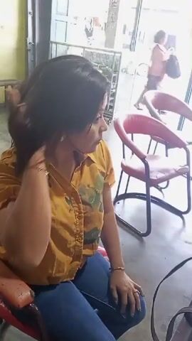 A woman with dark hair is sitting in a red chair. She is wearing a yellow collared shirt with a pattern on it and blue jeans. She is touching her hair and neck. There are pink chairs in the background.