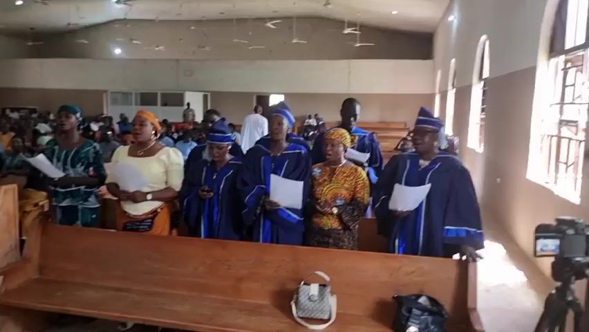 The video shows a wide shot of the interior of a church, filled with a large congregation. In the foreground, a group of people, likely a choir or religious leaders, stand on a raised platform or area, facing the congregation. They are wearing matching blue robes with white trim and some have academic-style caps. Several of them are holding papers, suggesting they are reading or singing from sheet music.

The congregation is seated in wooden pews, filling the majority of the church space. Many members of the congregation are also wearing traditional African attire with vibrant colors and patterns. The church itself appears to be a large, somewhat sparsely decorated hall with a high ceiling and exposed beams. Natural light streams in from arched windows on the right side of the frame.

In the immediate foreground, a wooden bench is visible, with a few handbags placed on it. A camera on a tripod is positioned to the right, indicating that the event is being filmed or recorded. The overall atmosphere appears to be one of a religious service or gathering.