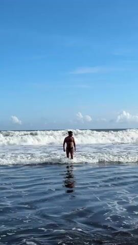 A single man is standing in the ocean. He is wearing a small black bathing suit and has his hair tied up on top of his head. He is facing towards the camera. Behind him are waves breaking in the ocean. Above the water is a blue sky with some white clouds.