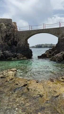 A video captures a view of a stone arch bridge connecting two rocky outcrops over a clear, greenish-blue body of water. The water appears shallow near the foreground, with visible rocks and ripples. In the distance, buildings and a hazy shoreline are visible. The sky is cloudy. The bridge has a stone archway and a railing made of rope and posts. No people are visible in the frame. The camera angle is low, looking up towards the bridge.