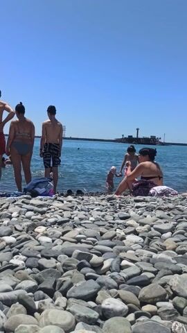 This is a short video clip of a crowded pebble beach on a sunny day. Several people are visible, some are wading in the shallow water, while others are sitting or standing on the beach. The people are mostly dressed in swimwear, suggesting a recreational activity like swimming or sunbathing.

In the foreground, a person in a black swimsuit and a red headscarf is holding a phone. Further down the beach, another person in a blue swimsuit is standing near the water. Several children are also present, playing near the shore. The sea is calm and blue, reflecting the clear sky. In the background, there are some industrial structures and a lighthouse, which might indicate the presence of a port or a coastal town. The overall atmosphere appears to be relaxed and cheerful, with people enjoying a summer day at the beach.

The time of day appears to be midday, given the bright sunlight and clear skies. The weather is warm and sunny, ideal for beach activities. The location is a pebble beach, common in the Black Sea region of Russia, specifically Bolshoy Sochi, as indicated by the presence of Olympic rings in the background and the general landscape. There is no visible text in the video.