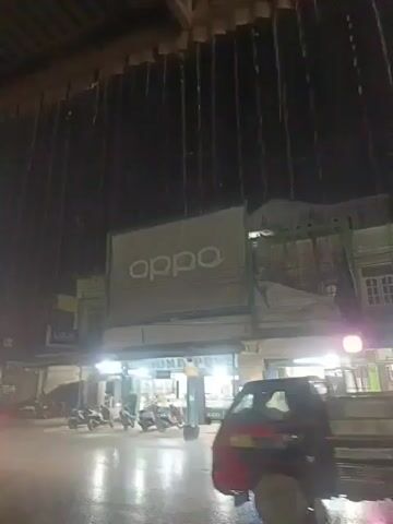 The video shows a street scene at night, possibly during or after rainfall. Water is dripping from above, creating streaks against the dark sky. In the background, there is a building with a large "oppo" sign displayed prominently. Below the sign, there are storefronts illuminated by lights. Several motorcycles are parked in front of the shops. A truck is visible on the street, and some people are riding on a motorcycle in the foreground. The road appears wet, reflecting the lights.
