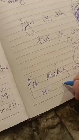 A person's hand is writing in a notebook with blue ink. The notebook is lined, and the writing is in cursive. The visible text includes "life do, did", "but yo should", "you", and "for writing all i'm so so". There is also a drawing of a smiley face and a scribbled circle in blue ink. The background appears to be a wooden surface.