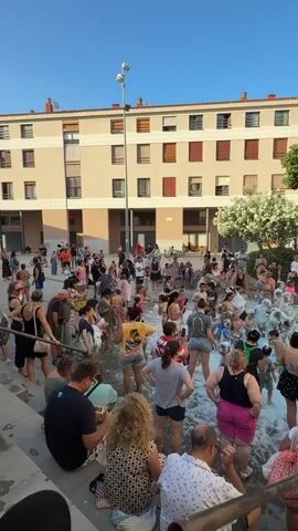 The video shows a daytime outdoor event with a large crowd of people gathered in what appears to be a plaza or courtyard. A building with multiple windows and balconies forms the backdrop. In the center of the gathering, a foam machine is spraying a large amount of white foam into the air, creating a foamy area where many people are interacting, dancing, and playing. Spectators are positioned around the perimeter of the foamy zone, some seated on steps. On the right side of the frame, there is a raised platform or stage with speakers and what look like bottles of foam solution. The sky is clear and blue, suggesting fair weather. People of various ages are present, many dressed in summer attire like shorts and tank tops. The overall atmosphere appears festive and lively.