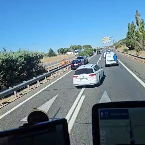 The video shows a highway scene with several cars stopped on the shoulder and in a lane. Two individuals wearing orange vests are visible near a black car, and appear to be interacting with its occupants or the vehicle itself. A white car is stopped behind the black car, followed by a grey SUV. Further down the road, a blue and white vehicle, possibly a police car or service vehicle, is also stopped. A large truck is visible in the distance in the leftmost lane. The environment appears to be rural with dry vegetation and a clear blue sky. The perspective is from inside a vehicle, looking out through the windshield, with two navigation devices visible in the foreground. The overall impression is a roadside incident or breakdown.