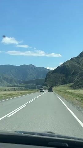 The video shows a view from inside a vehicle driving on a highway. The highway is paved and has white lines marking the lanes. Two cars are visible ahead on the highway, driving in the same direction as the viewer's vehicle. To the right of the highway, there is a grassy slope leading up to a large, green, mountainous area. The mountains are covered in vegetation and appear to be covered in shadows in some areas. In the distance, more mountains are visible, appearing layered and less green. The sky is blue with some scattered white clouds. The overall scene depicts a scenic drive through a mountainous landscape on a sunny day.