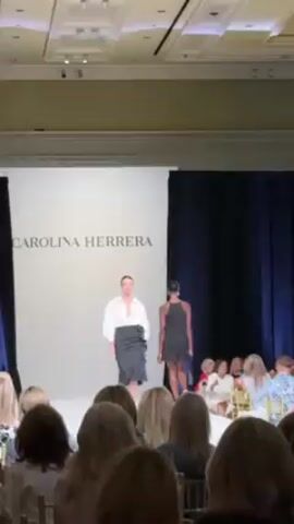 The video shows a fashion show, with a model walking down the runway wearing a white blouse and a black skirt. Behind her, the name "CAROLINA HERRERA" is displayed on a white background.  Another model, in a black dress, is walking behind.  The audience, consisting mostly of women, is seated in the foreground. The runway is well-lit, and dark blue curtains are visible on the right side. The event is taking place indoors.