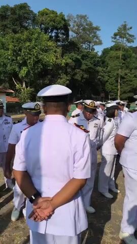 A group of uniformed individuals, mostly men in white naval uniforms with hats, are standing outside on a clear day. The background consists of lush green trees and a bright blue sky. Some individuals are looking down or adjusting their uniforms, while others appear to be listening or conversing. The ground is covered with dry grass and dirt. The lighting suggests it is daytime.