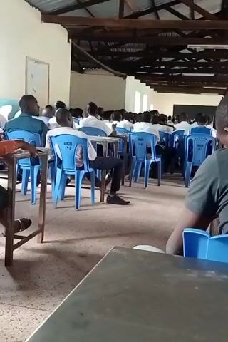 The video captures a large gathering of people, predominantly young men, seated in rows within a spacious hall or classroom. The setting features a high, exposed wooden truss ceiling with fluorescent lighting and light-colored walls. Blue plastic chairs are arranged in front of wooden desks, with most attendees facing forward.

At the front of the room, a person stands, seemingly addressing the audience, with blackboards visible in the background. The attendees appear to be listening attentively, with some looking down, possibly taking notes or focused on the speaker. Natural light enters through windows along the side walls, complemented by artificial lighting.

Notable details include white text, "IBORA 112," visible on the back of one of the blue plastic chairs. The overall scene suggests an educational session, lecture, or community meeting in Lwandeti ward, Kenya, taking place during the daytime. The atmosphere appears calm and focused.