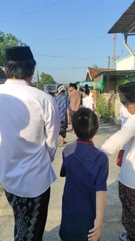 A group of males walks down a street on a clear day. The group comprises men and boys, some wearing traditional Muslim attire, including white shirts, black caps, and patterned sarongs. They walk along a street lined with houses and electric poles. People on motorcycles and on foot occupy the street. One man looks at his mobile phone. The atmosphere suggests a communal gathering or procession, possibly related to a religious event.