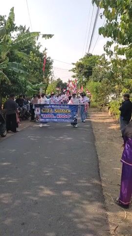 This is a vertical, outdoor, eye-level shot of a parade or procession. A group of people, predominantly young, are walking in single file down a paved road. They are mostly dressed in white shirts and some are wearing masks. At the forefront of the procession, several individuals hold a large banner that reads "CITA SUARA" and "LLOO. KOGG." The banner is blue with white text and some graphics. The participants are holding small Indonesian flags. Spectators are visible on the left side of the road, some sitting and some standing. There is also a motorcycle parked on the left. Trees and vegetation line both sides of the road, and utility poles with wires are visible in the background. The sky is clear and the lighting suggests it is daytime, likely late afternoon due to the long shadows.