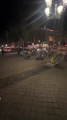 The video depicts an outdoor scene at night. The ground is covered in cobblestones, and a gray concrete bench is visible in the foreground. Several parked bicycles lean against each other, and a few are lying on the ground. A street lined with buildings stretches into the background, some illuminated with vibrant green, red, and white lights. Cars drive on the street, and a police car with flashing lights is visible. People can be seen standing around, and streetlights provide additional illumination.