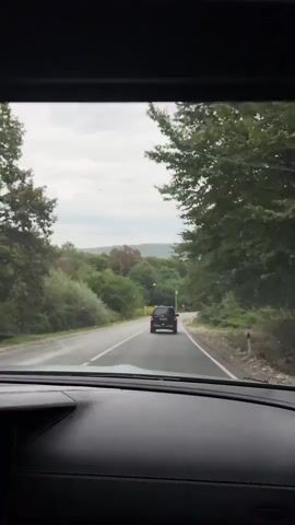 The video shows a view from inside a car looking through the windshield. The car is driving on a paved road that winds through a heavily treed area. In front of the car, another dark-colored SUV is driving ahead. The sky is overcast, and the overall lighting suggests it is daytime. The road has white lines marking the lanes and has a shoulder on the right side with some gravel and vegetation. There are trees and foliage on both sides of the road, with some trees also visible in the distance on rolling hills. The dashboard of the car is visible in the foreground, suggesting the footage is captured from the driver's or passenger's perspective.