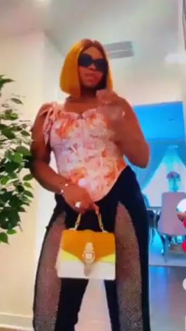 A woman is standing and dancing in what appears to be a hallway or living room. She is wearing sunglasses, a patterned top, black pants with a see-through mesh or sparkly design down the sides, and holding a two-toned white and yellow purse. There is a green plant to the left and a glimpse of a dining area in the background. The woman is making hand gestures as she dances.