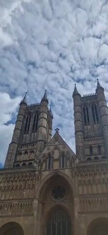 The video shows a low-angle view of the entrance and towers of Lincoln Cathedral against a cloudy sky. The cathedral is a large stone structure with intricate carvings and tall, pointed towers. The central entrance is framed by an archway and windows above. The towers are composed of multiple levels with windows and spires.  The sky is filled with puffy white clouds against a blue backdrop. Church bells ring in the background.
