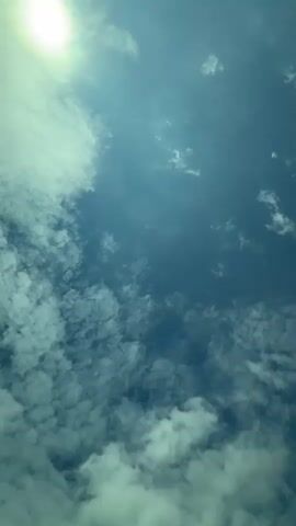 The video shows a view of a bright sun in a light blue sky with many small, fluffy white clouds. The camera is pointed directly upward. The light is bright, making the clouds appear almost white and the sky a deeper blue.