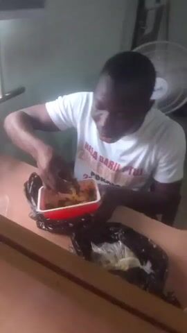 A young Black man wearing a white t-shirt with text on it is sitting at a wooden table and eating from a red plastic container. He is using his fingers to scoop food from the container and bring it to his mouth. Next to the red container is a black plastic bag with some white items inside. In the background, there is a wall, a fan, and a window. The lighting in the room appears to be artificial.