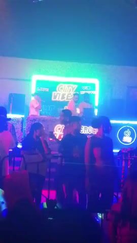 The video captures an energetic indoor nightclub or bar scene at night, brightly illuminated by vibrant neon lights in shades of blue, green, pink, and purple. Numerous individuals are gathered, creating a lively atmosphere. At the center, a raised stage or DJ booth features two people behind what appears to be DJ equipment, likely performing or mixing music.

Behind the performers, a prominent large screen or LED sign displays "City Vibes" in glowing green neon. Faintly visible text above "City Vibes" includes "Life" and "UB". Below the main stage, another neon sign shows "City VIBES" and "CROCITY". Speakers are positioned on either side of the stage setup. The audio includes music and a male voice, likely a DJ or host, speaking to the crowd, suggesting an active event. The setting is indicative of a nightlife venue in Rido, Nigeria.