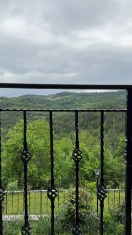 The video shows a view of a lush, green forest landscape from a balcony. The camera is positioned behind a black wrought iron railing with decorative twists. The sky is overcast with gray clouds. In the distance, there are rolling hills covered in trees, with a small village visible atop one of the hills. Below the balcony, there's a well-maintained lawn with various plants and a single lamppost. The camera slowly moves to the right, revealing the wooden frame of the balcony.