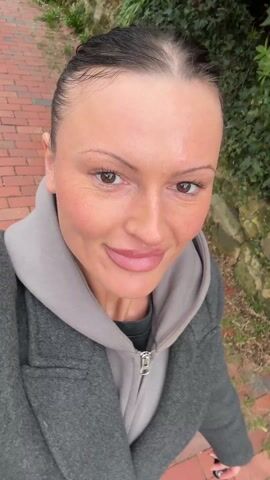 A smiling woman with brown eyes and neatly pulled-back dark hair looks directly at the camera. She is outdoors on a red brick path, wearing a gray coat over a light gray hoodie. The camera appears to be held by the woman, capturing a high-angle selfie perspective. Behind her, there is greenery.